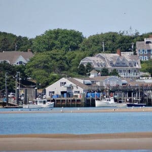 Chatham harbor from the beach facing the town fish pier with fishing boats, large pier with shingled buiding and stately cape cod style homes in background surrounding by tall green trees.