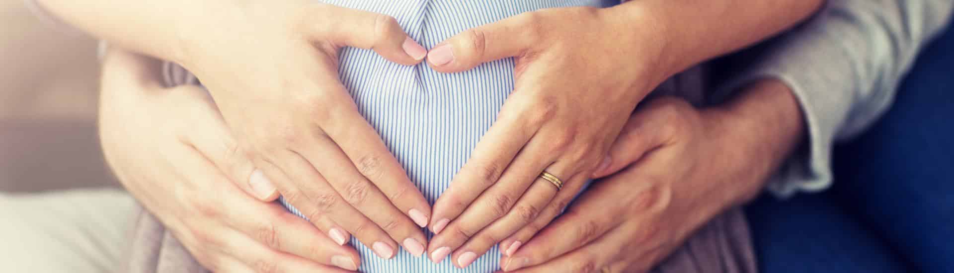 Woman making a heart shape with her hands over her stomach with a mans hands below