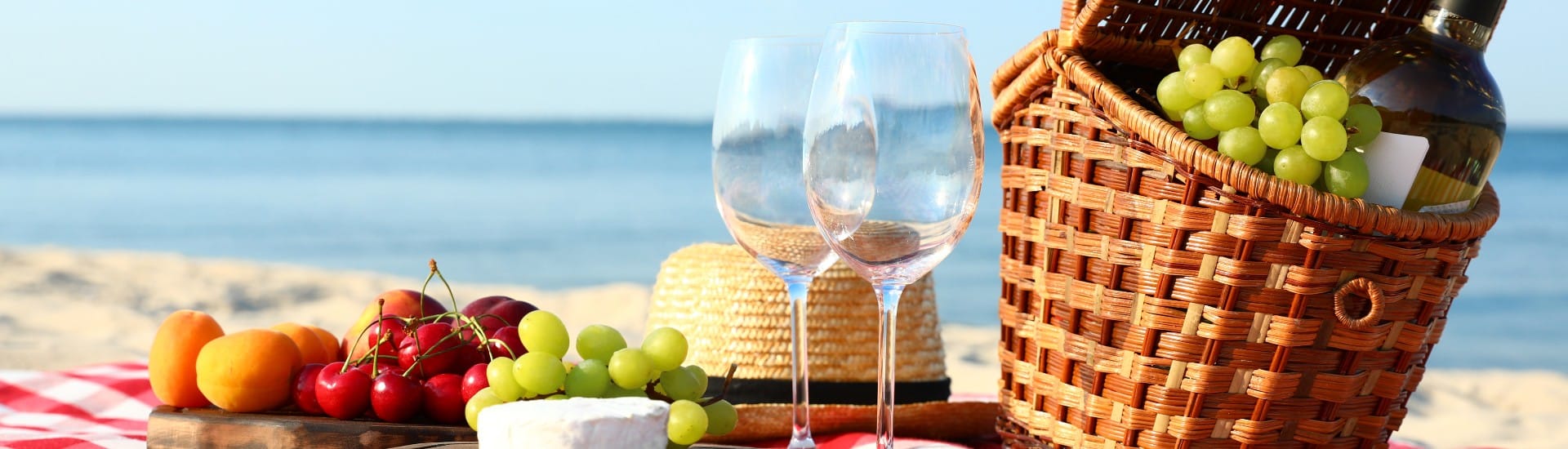 Brown wicker picnic basket with a hat, wine glasses and fruit on a wood platter