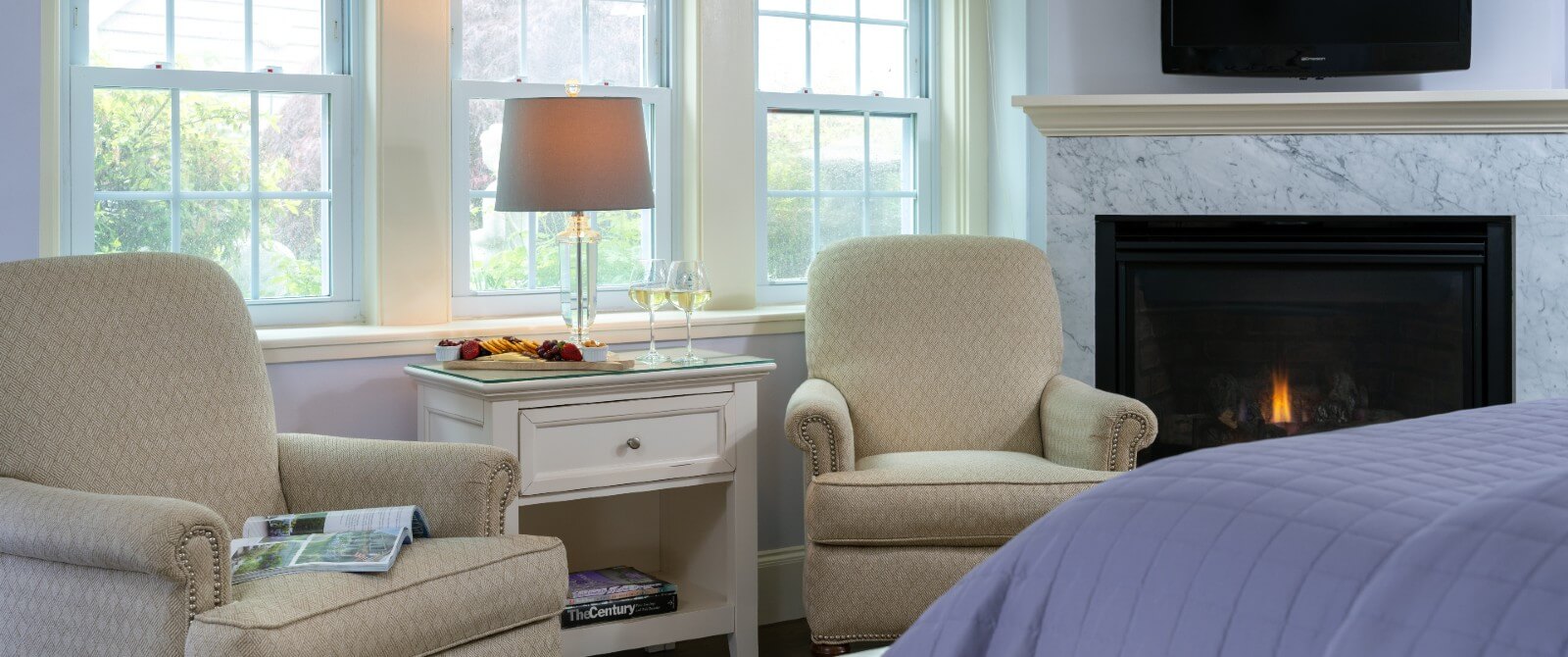 Two beige sitting chairs with table holding lamp and fruit tray next to a gas fireplace