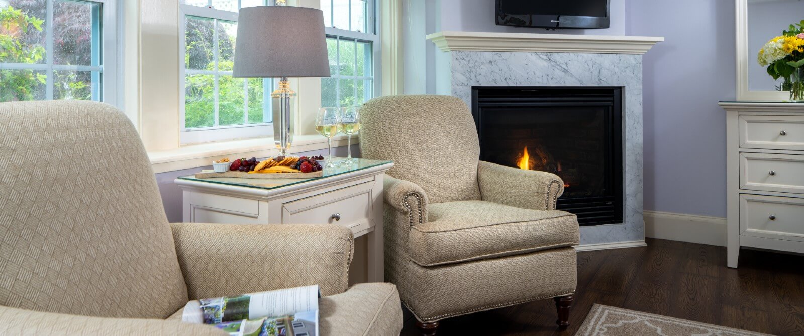 Two beige sitting chairs with table holding lamp and fruit tray next to a gas fireplace