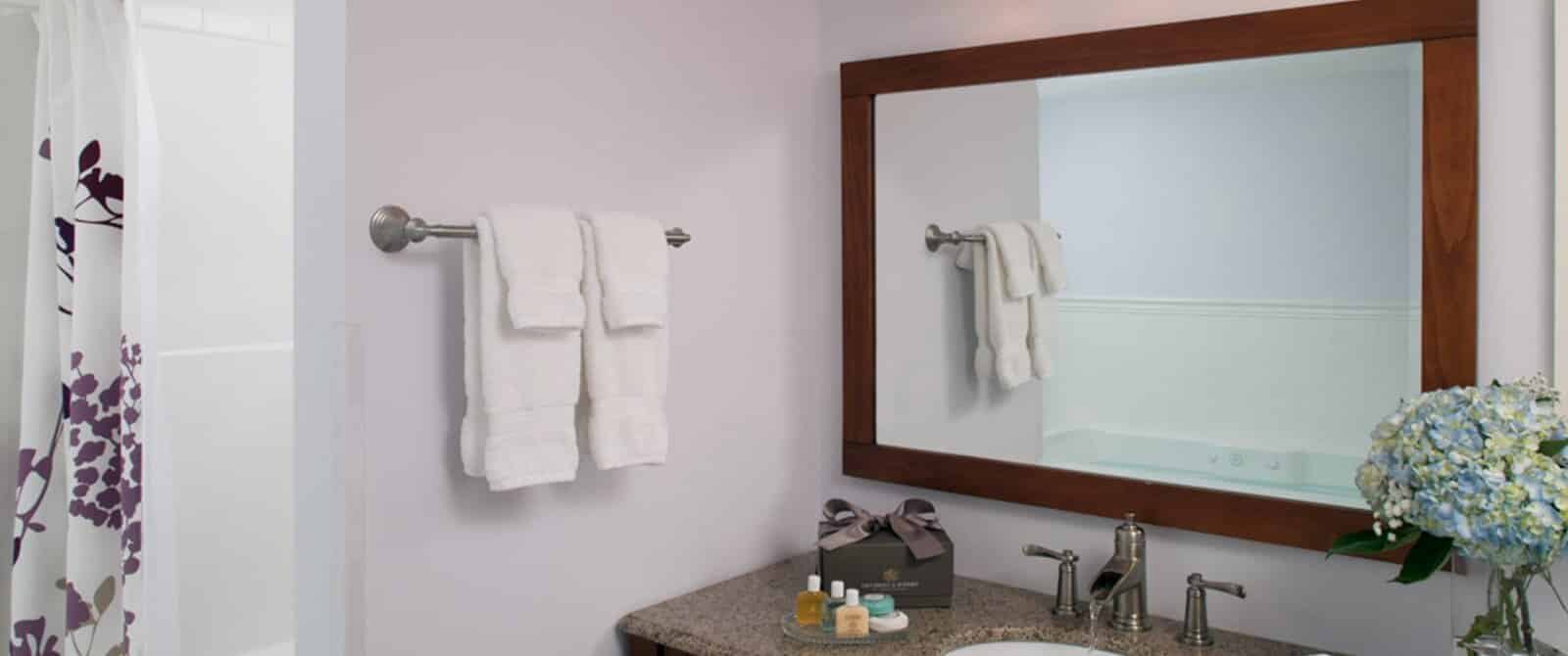 Bathroom with single sink, pale lilac walls, hanging white towels and large wood-framed mirror