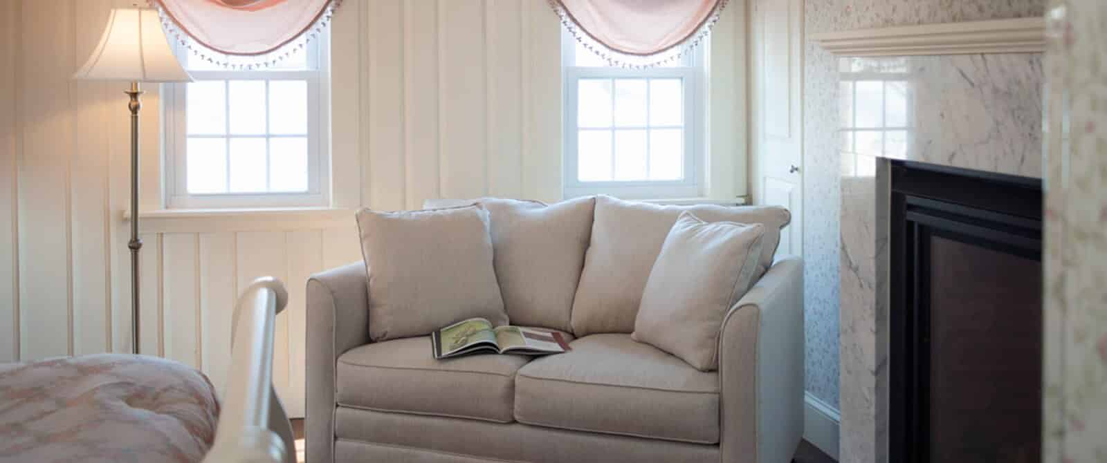 Beige loveseat in the corner of a bedroom by two windows and fireplace