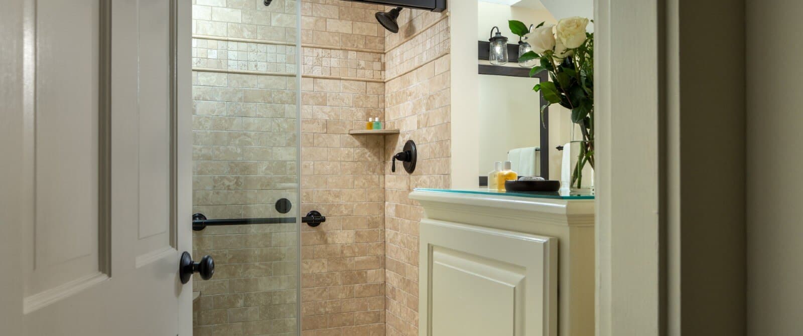 Open door to bathroom with tiled shower with glass doors and cabinet with vase of flowers