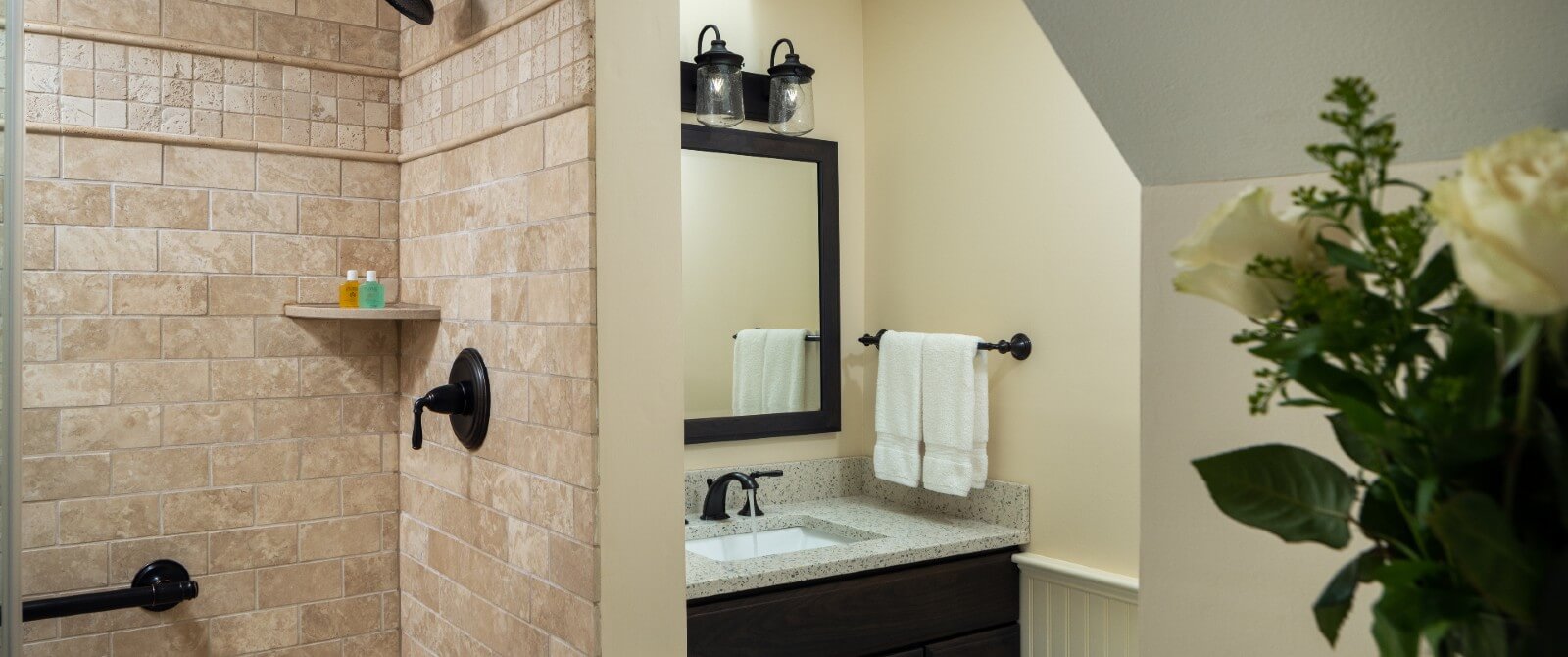 Shower with brown tile and glass doors by sink vanity under framed mirror