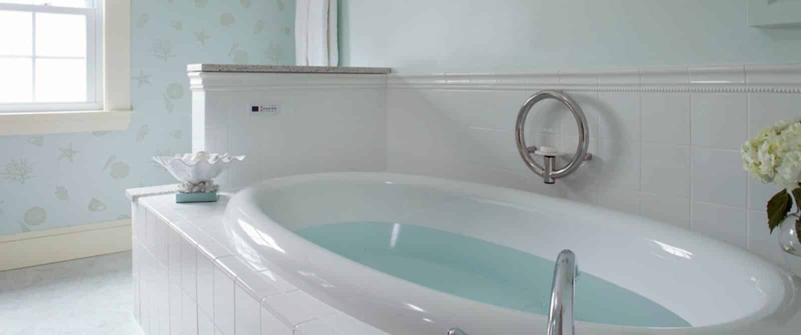 Large oval soaker tub filled with water in bathroom with white tile bright window
