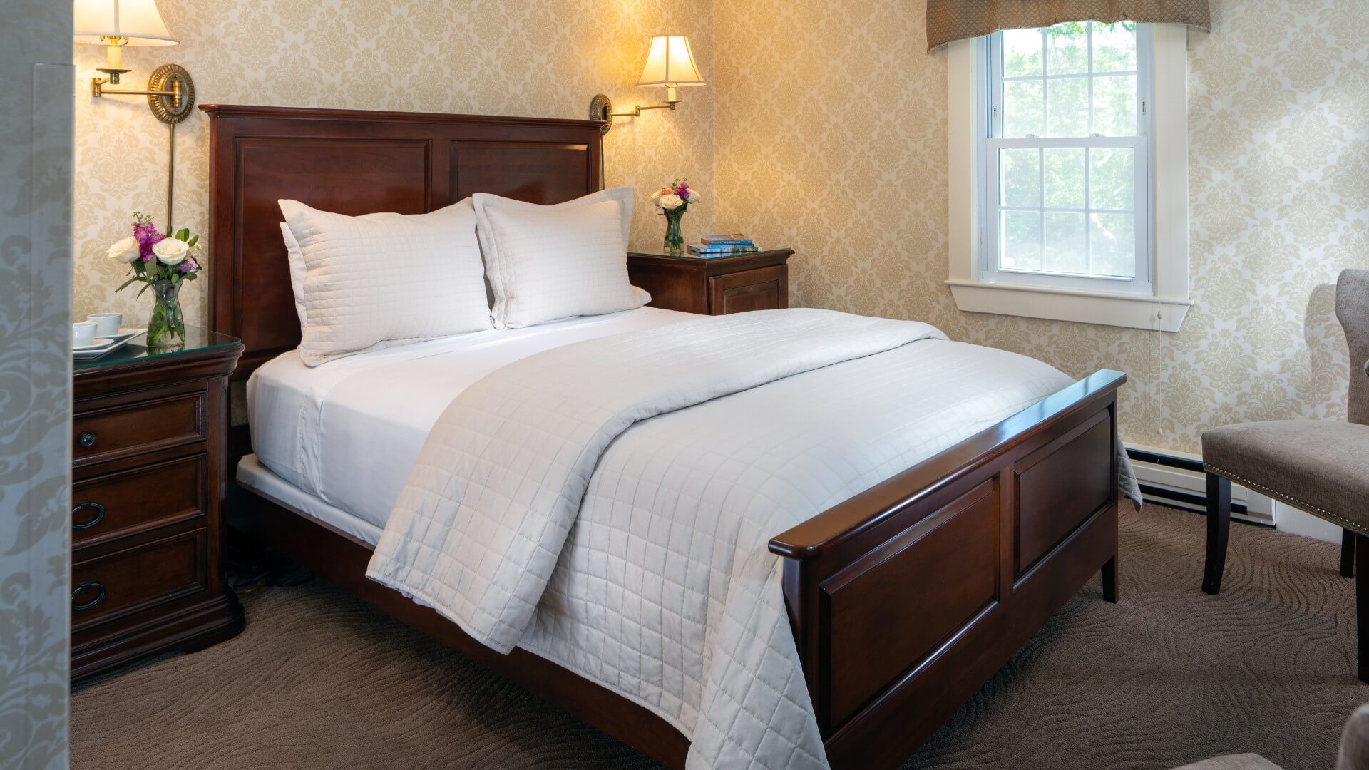 Elegant bedroom with bed in white quilt, decorative wallpaper and sitting chair by a window
