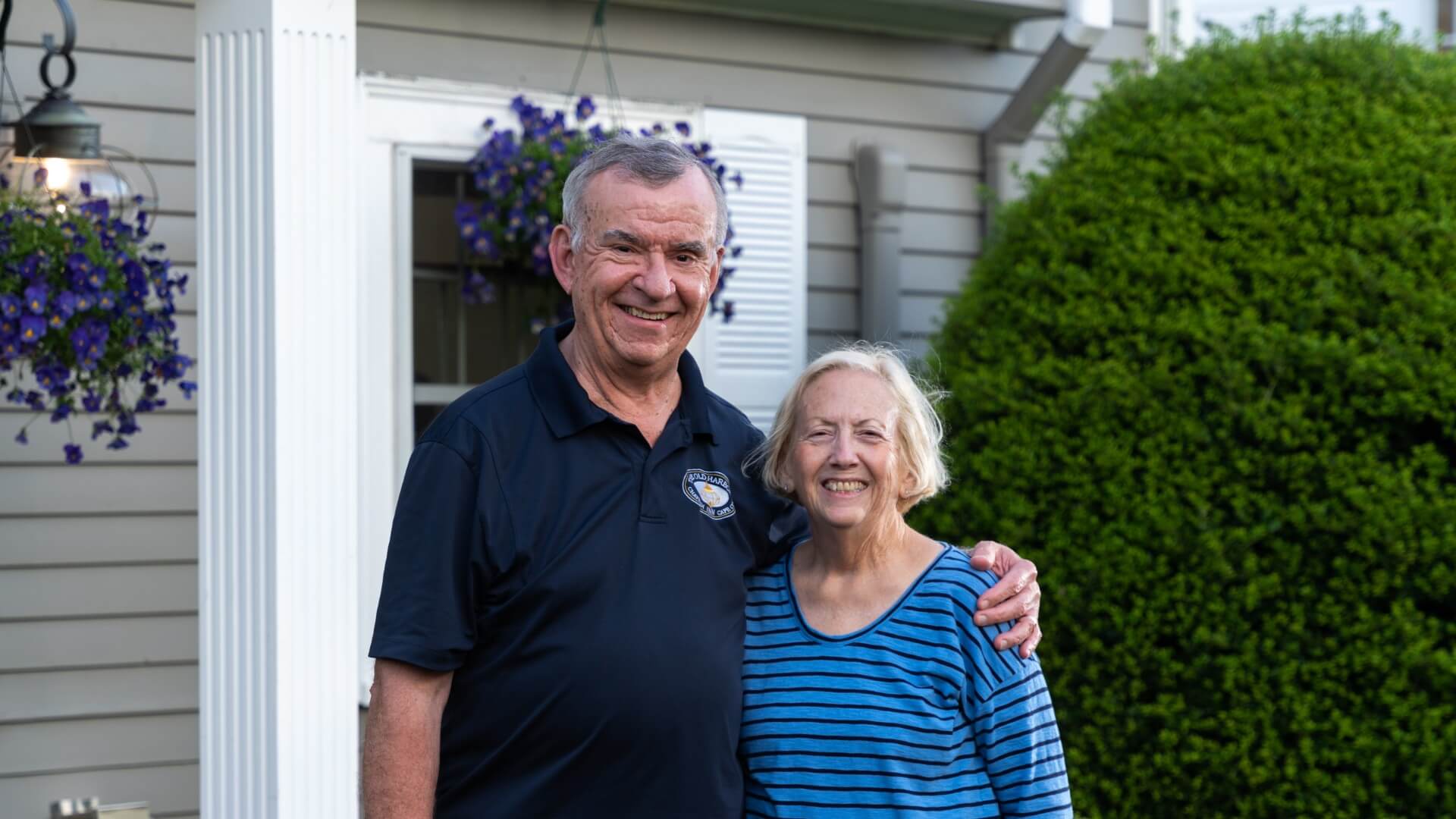 A man and woman standing together smiling in front of a home with flowers and green bushes