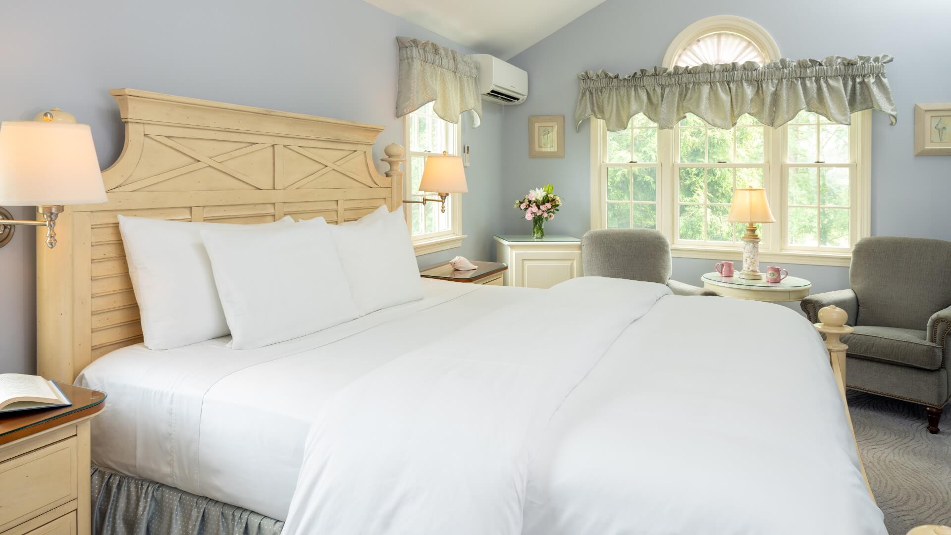 Spacious bedroom with king bed in white linens, and two sitting chairs under a bright window