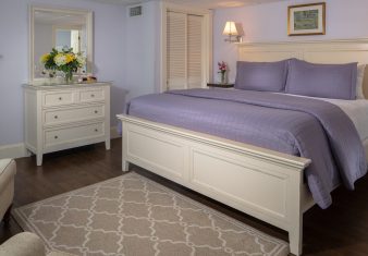 Spacious bedroom in purple hues with king bed, dresser with mirror, fireplace and two sitting chairs
