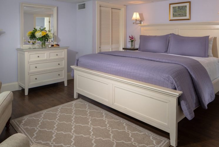 Spacious bedroom in purple hues with king bed, dresser with mirror, fireplace and two sitting chairs