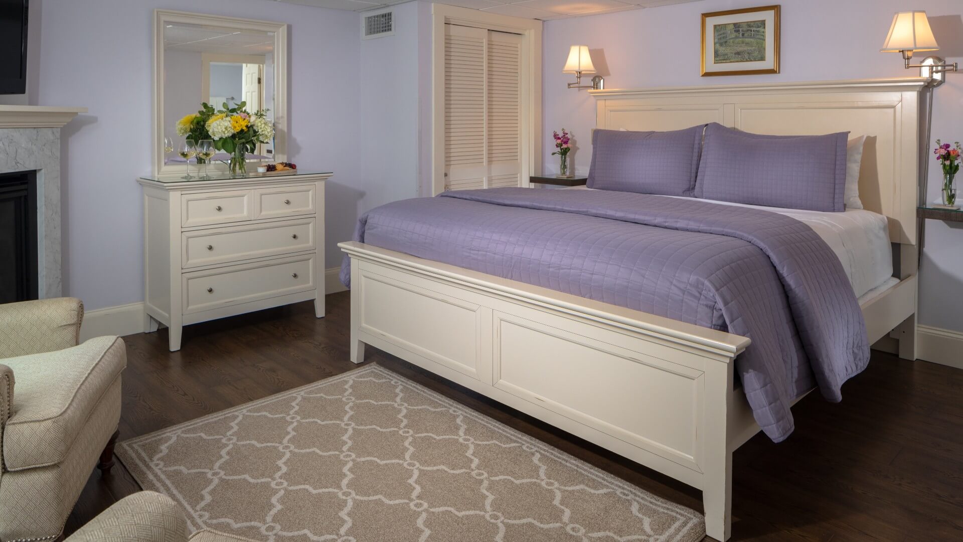 Spacious bedroom in purple hues with king bed, dresser with mirror, fireplace and two sitting chairs