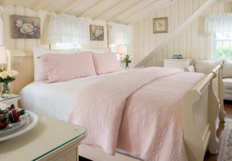 Bright bedroom with queen bed with pink quilt, slanted ceilings, loveseat and table with wine and strawberries