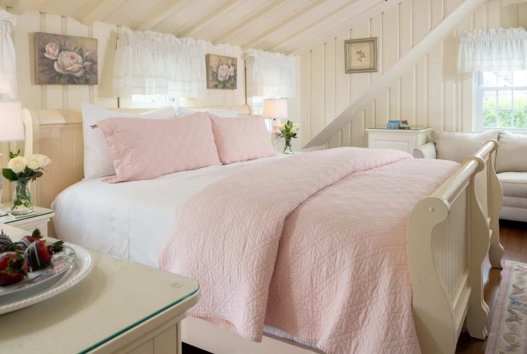 Bright bedroom with queen bed with pink quilt, slanted ceilings, loveseat and table with wine and strawberries