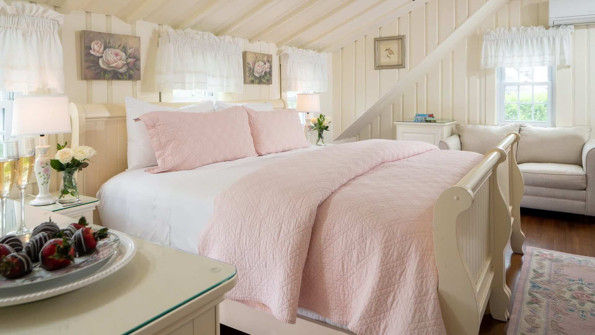 Bright bedroom with queen bed with pink quilt, slanted ceilings, loveseat and table with wine and strawberries