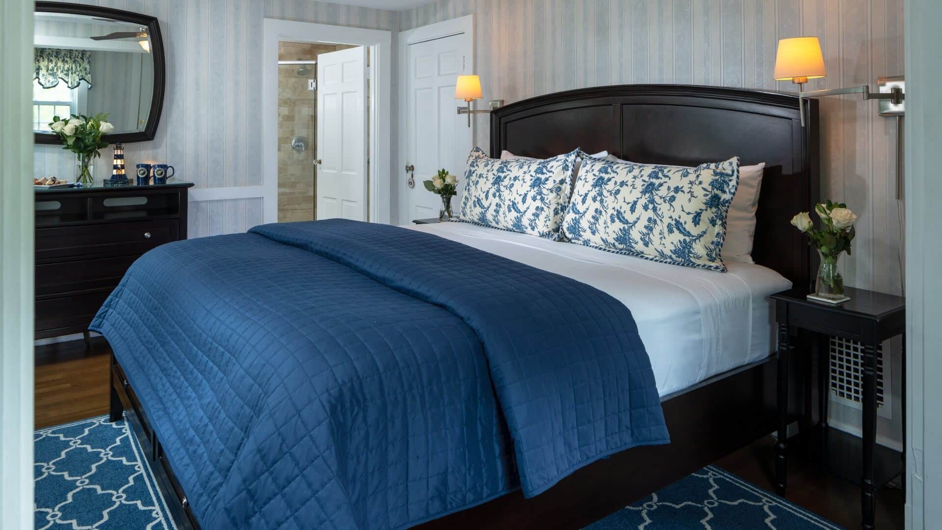 Elegant bedroom in blue hues with king bed, dresser with mirror and doorway into attached bathroom