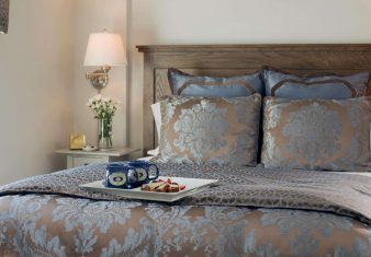 Queen bed in decorative comforter with tray of coffee mugs and baked goods in a bright bedroom