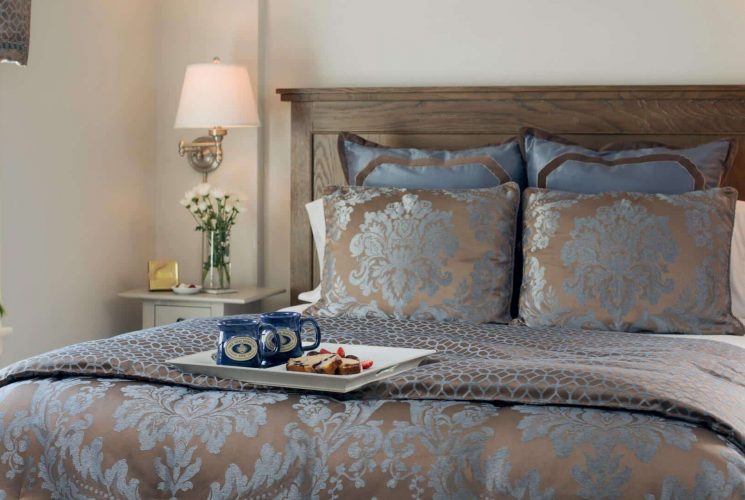 Queen bed in decorative comforter with tray of coffee mugs and baked goods in a bright bedroom