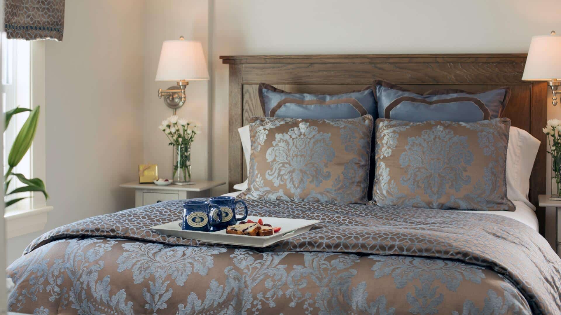 Queen bed in decorative comforter with tray of coffee mugs and baked goods in a bright bedroom