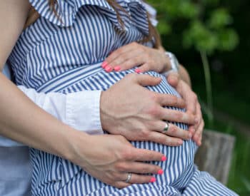 Pregnant woman wearing blue and white striped outfit embraced by a man whose hands are holding her belly