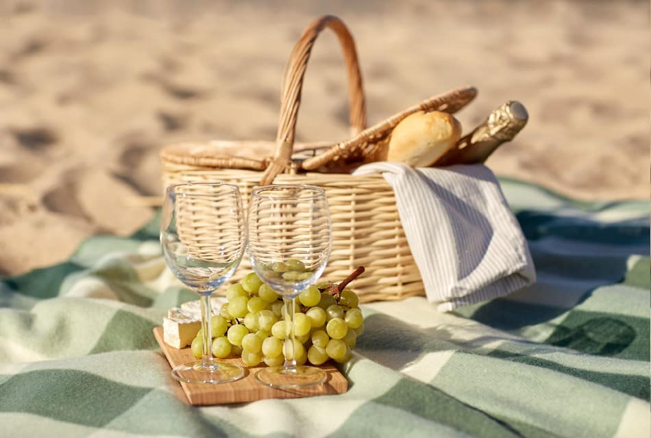 Picnic Basket filled with french baguette and bottle of sparkling wine and next to it a cutting board with grapes and cheese on top of a cozy blanket