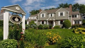 Front facade of a large home with lush landscaping and flowers, green lawn and business sign on white posts