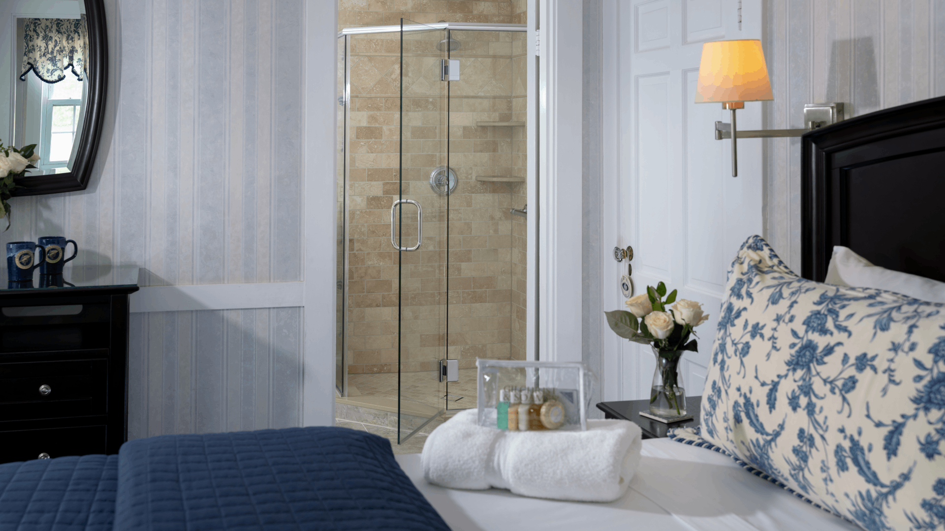 Bedroom with bed with white and navy linens, towels on the bed fresh flowers and view of the bathroom