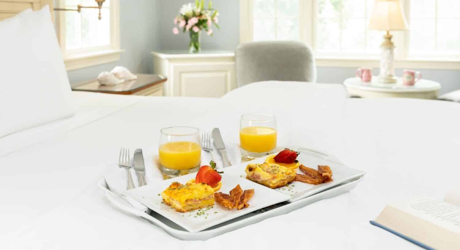 Breakfast tray with two plates of food, two glasses of orange juice and silverware on a bed with white linens