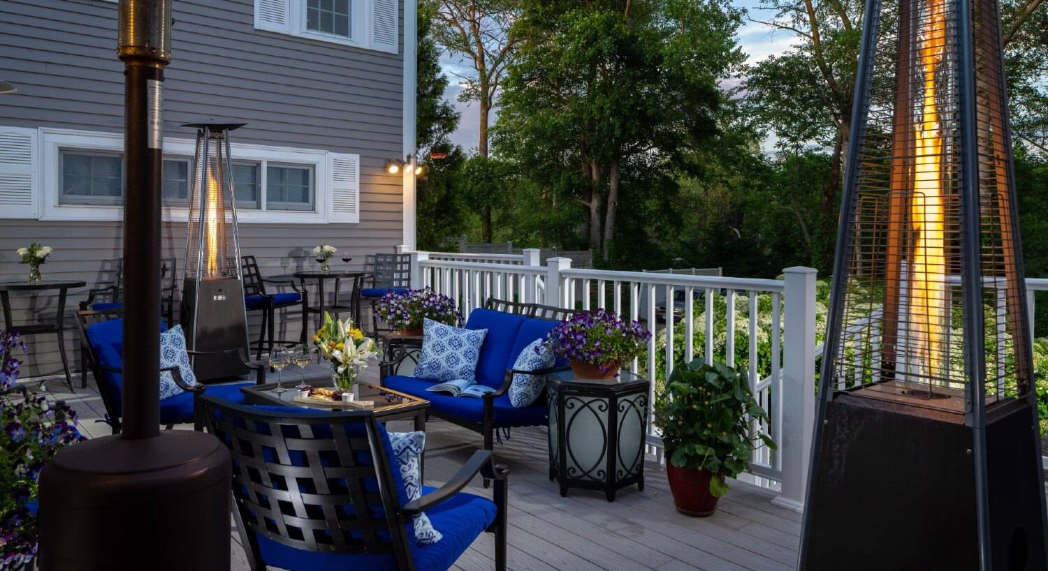 Outdoor deck at dusk with patio furniture, potted flowers and tall fire lamps