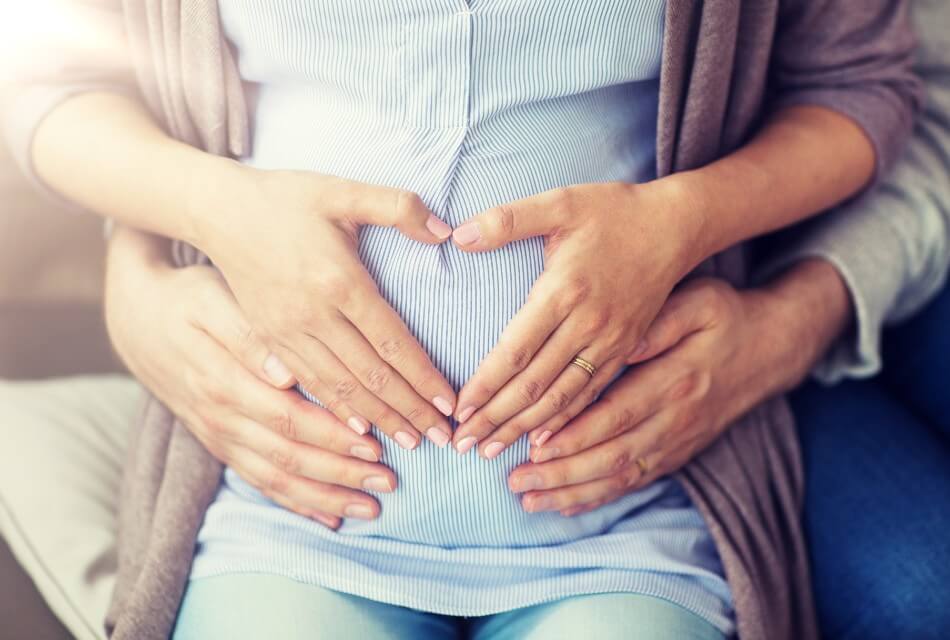 A woman making a heart shape with her hands over her belly with a mans hands below