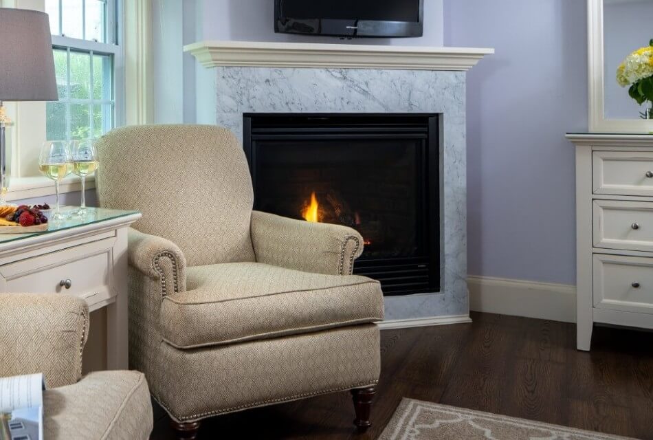 Beige sitting chairs with table holding wine and fruit by a gas fireplace