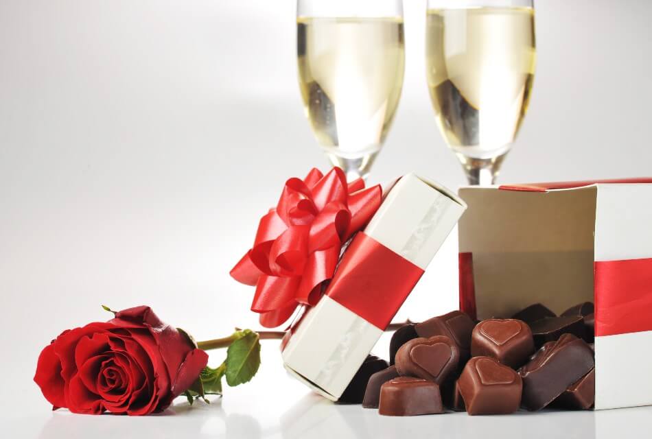 Two champagne glasses by a tipped over box of chocolates and a red rose