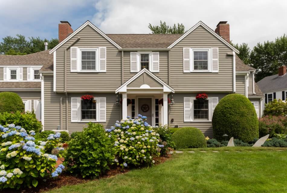 Front facade of a home with brown siding, white shutters and large hydrangea bushes