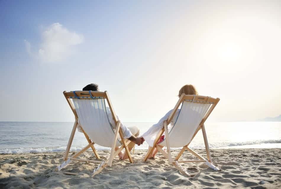 A man and woman in white beach chairs sitting in the sand by waters edge holding hands